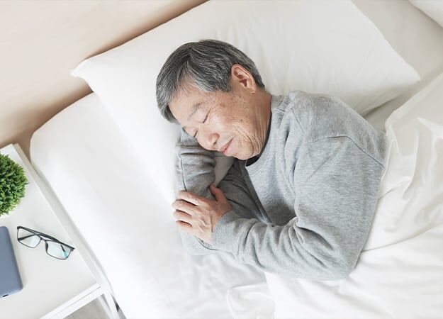 12Oaks-Tampilan atas pria senior Asia berbaring di tempat tidur, tidur nyenyak dengan senyum di kamar tidur di rumah-ss-Posisi Tidur Terbaik untuk Lansia